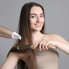Uśmiechnięta kobieta z długimi brązowymi włosami suszy je suszarką i szczotką, tworząc idealny styling włosów.