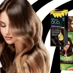 Kobieta z pięknymi, falowanymi włosami w odcieniu popielaty brąz. Obok opakowania farb do włosów Garnier Olia, Joanna i Wella.