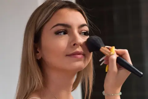 Młoda dziewczyna z długimi blond włosami nakłada puder pędzlem. Jej makijaż jest subtelny, podkreślający naturalne piękno, w stylu "clean girl".