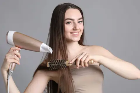 Uśmiechnięta kobieta z długimi brązowymi włosami suszy je suszarką i szczotką, tworząc idealny styling włosów.