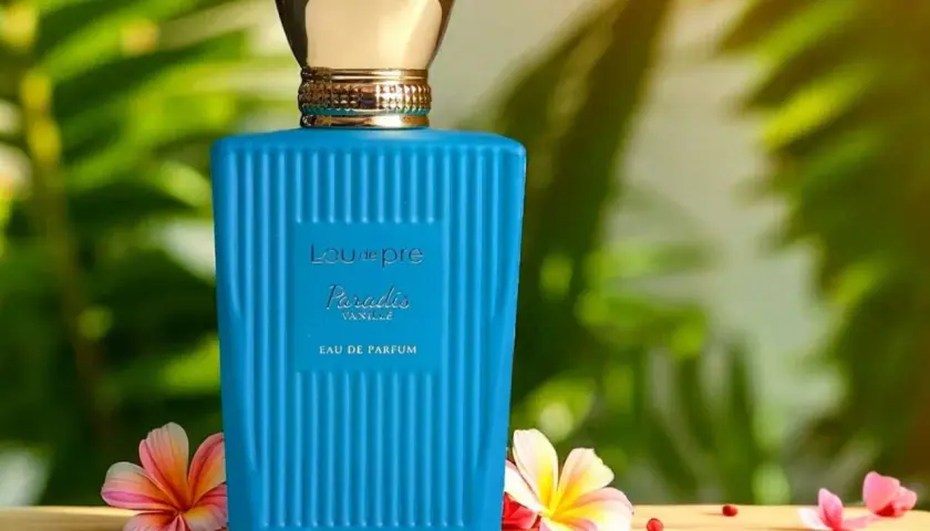 Błękitna butelka perfum Lou de Pre Paradis Vanille z złotą nakrętką, otoczona tropikalnymi kwiatami i liśćmi.