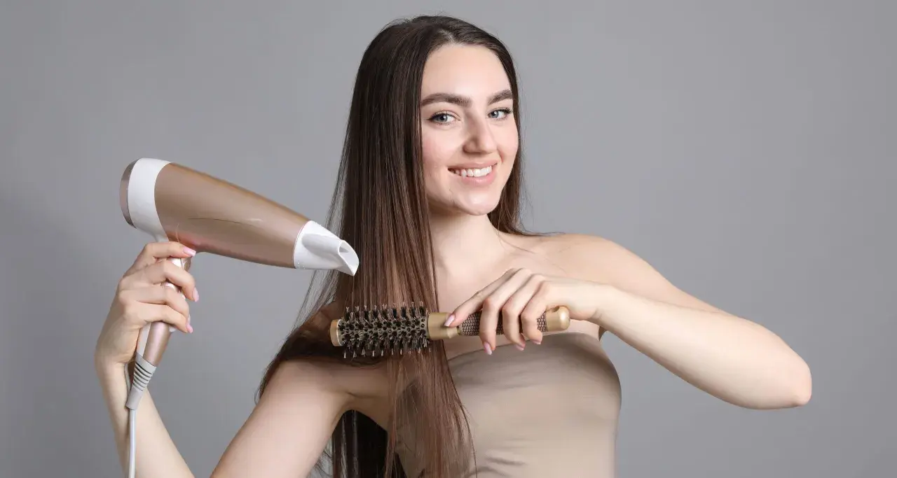 Uśmiechnięta kobieta z długimi brązowymi włosami suszy je suszarką i szczotką, tworząc idealny styling włosów.