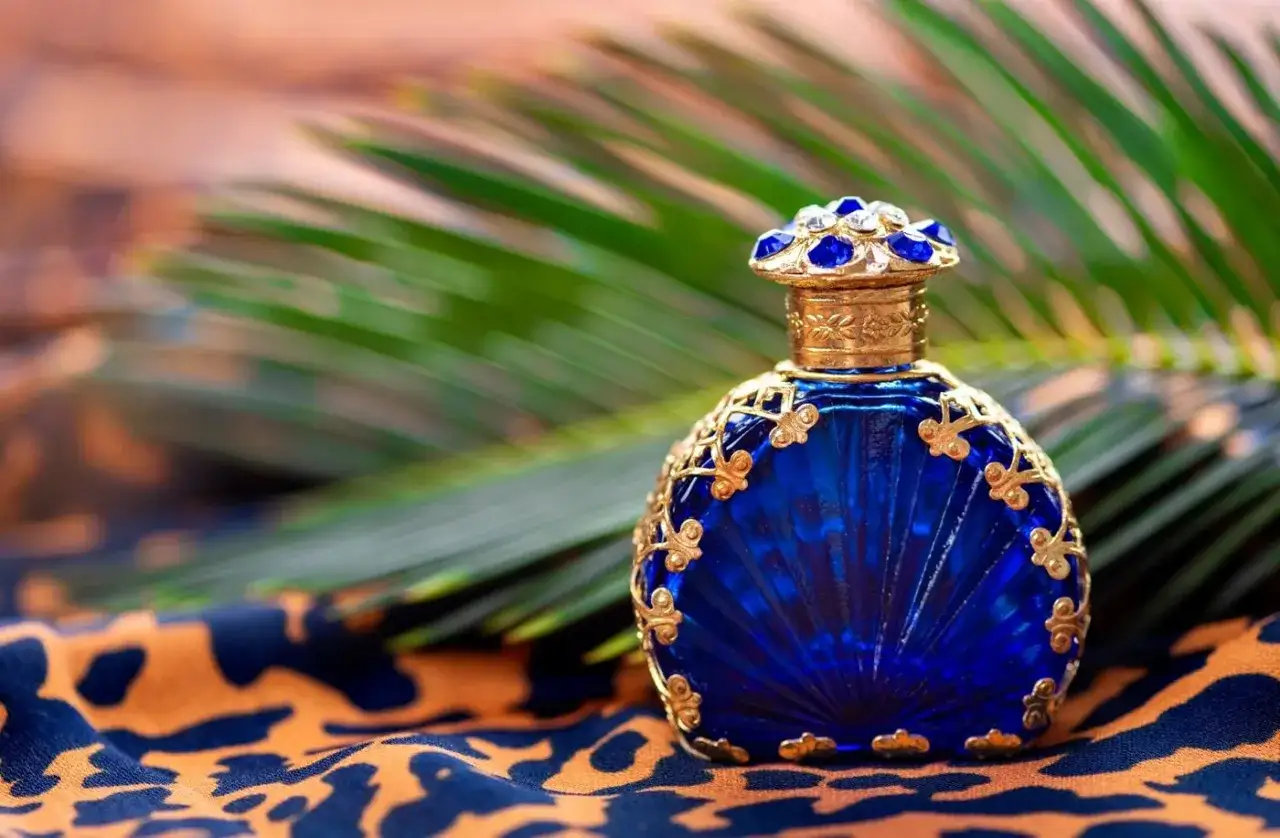 Elegancka, kobaltowa buteleczka perfum lou de pre, ozdobiona złotymi zdobieniami i szafirami, na tle liści i wzorzystej tkaniny.